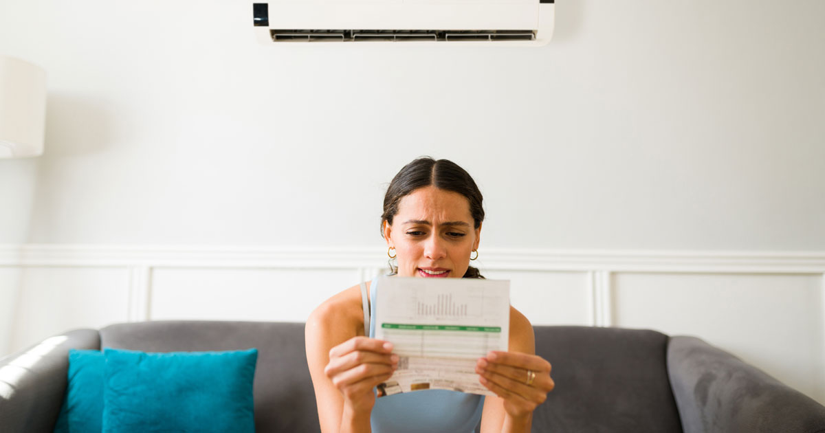 Femme assise sur un canapé regardant sa facture d’électricité avec inquiétude, sous un climatiseur mural en fonctionnement dans un salon moderne.
