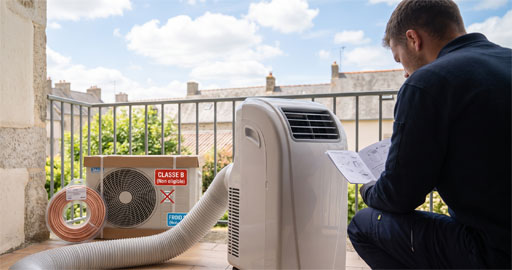 Un technicien, vu de dos, est accroupi sur un balcon et lit un manuel. À côté de lui se trouve un grand climatiseur mobile blanc avec son tuyau d'évacuation flexible. En arrière-plan, on voit l'unité extérieure d'un climatiseur split dans son carton d'emballage, portant l'étiquette rouge "Classe B (Non éligible)", ainsi qu'un rouleau de tube en cuivre. Le balcon donne sur les toits d'un village par une journée ensoleillée.